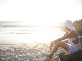 Frau mit weißem Sonnenhut sitzt am Strand und schaut ins Meer