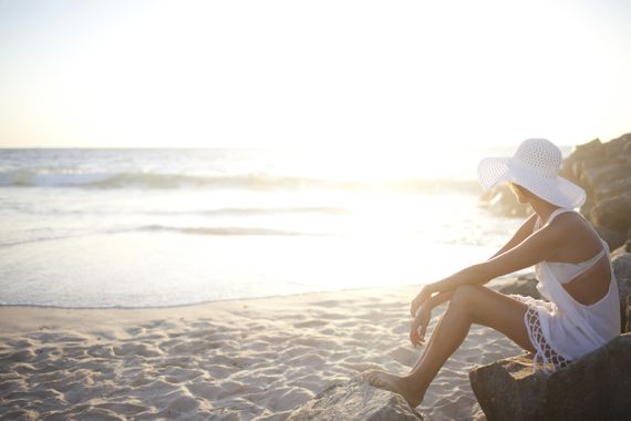 Frau mit weißem Sonnenhut sitzt am Strand und schaut ins Meer