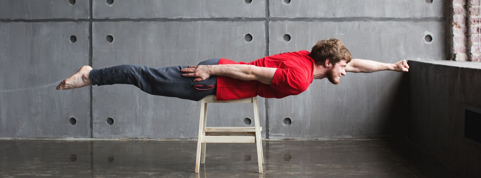 Junger Mann mit rotem T-Shirt streckt sich auf Hocker zur Fliegerpose aus